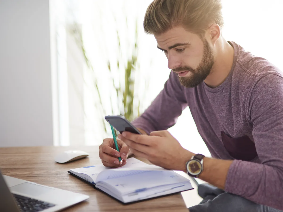 Mann rechnet am Schreibtisch mit Notizbuch und Handy die Kosten seiner MPU zusammen